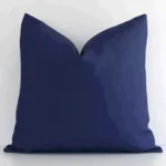 White wall with a solid colour navy cushion laying against it. It has a distinctive outdoor fabric and has a square shape.