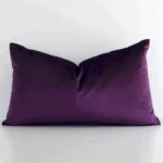 Ractangular solid-coloured cushion cover in plum purple colour sitting upright in front of a white wall. It has been made from a quality velvet material.