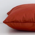 A square rust cushion cover laying flat. This viewpoint highlights the solid colour style and velvet fabric from a side position.