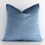 Stone blue solid-coloured cushion positioned in front of a white wall. It has square shape and is made from a velvet material.