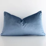 A lovely rectangular stone blue cushion cover arranged in front of a white wall. The solid colour style complements the velvet material.